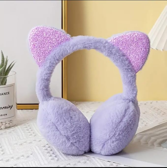 The image shows a pair of purple, fluffy earmuffs with cat ear designs featuring pink glitter accents, placed on a white textured surface. In the background, there is a gold-framed mirror, a small plant in a labelled jar, and a folded paper or cloth.
