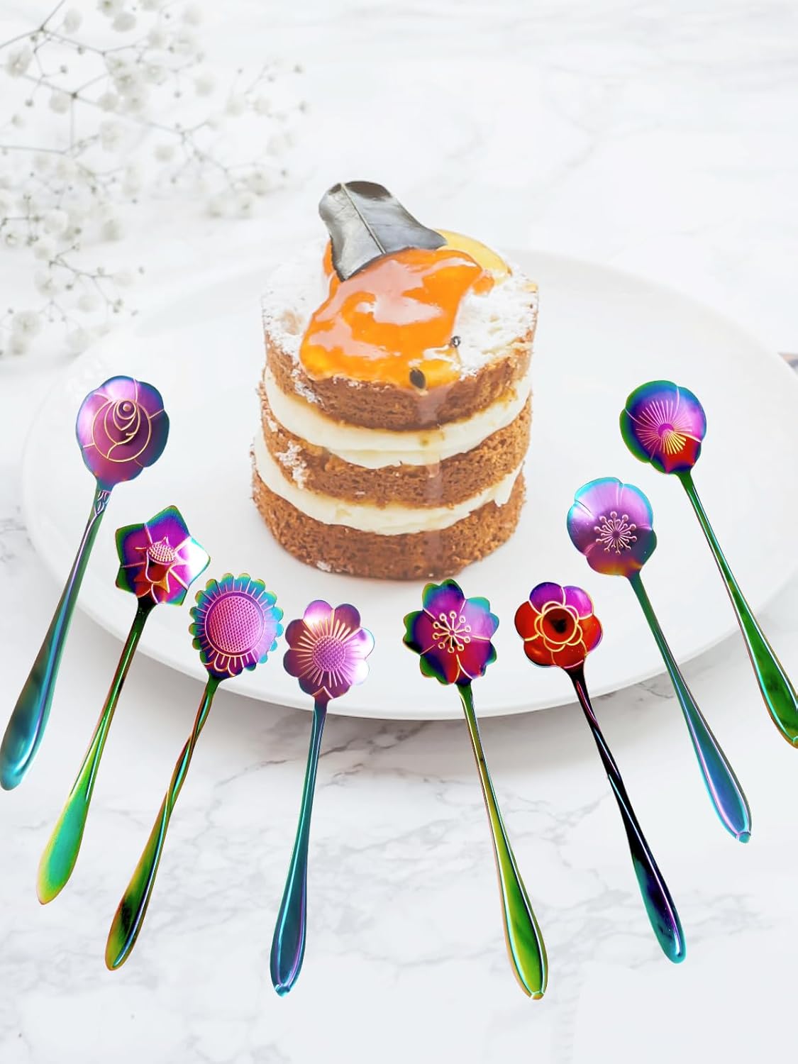 A small layered cake with orange glaze and cream sits on a white plate, surrounded by iridescent spoons with floral designs. The background features a marble surface and white flowers.