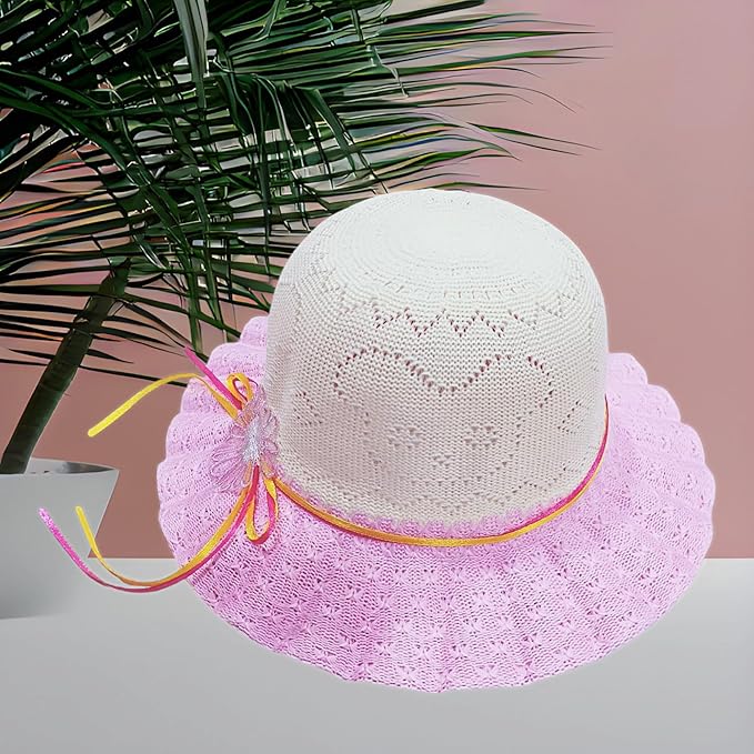 The image shows a white crocheted hat with a wide, pink lace brim, featuring a pink and yellow ribbon with a decorative bow, placed on a surface with a green plant and a pink background.