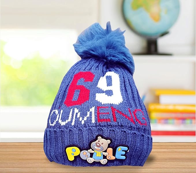A blue knitted baby hat with a fluffy pom-pom features red "6" and "9", white "OUMENC" text, and a colorful "Pooie" bear patch. Placed on a wooden surface, it’s set against a blurred background with a globe and colorful books, suggesting a kids' room.