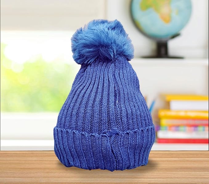 A blue knitted baby hat with a fluffy blue pom-pom sits on a wooden surface. The background features a blurred view of a globe and colorful books, suggesting a children's room setting.