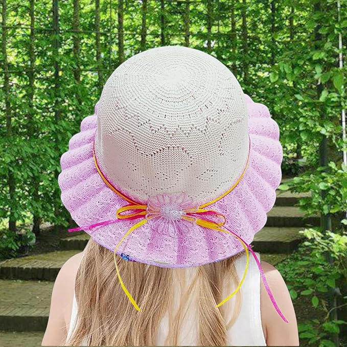 The image shows a child with long blonde hair wearing a white crocheted hat with a pink lace brim, tied with a pink and yellow ribbon, standing outdoors with a lush green background and stone steps.