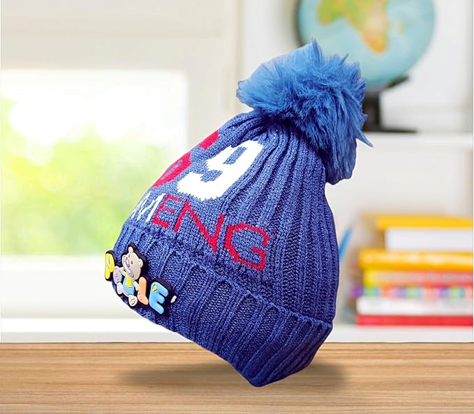 A blue knitted baby hat with a fluffy blue pom-pom features red "6" and "9" numbers and white "OUMENC" text. It has a colourful "Pooie" bear patch on the front. The hat rests on a wooden surface with a blurred background of a globe and colourful books, indicating a children's room.